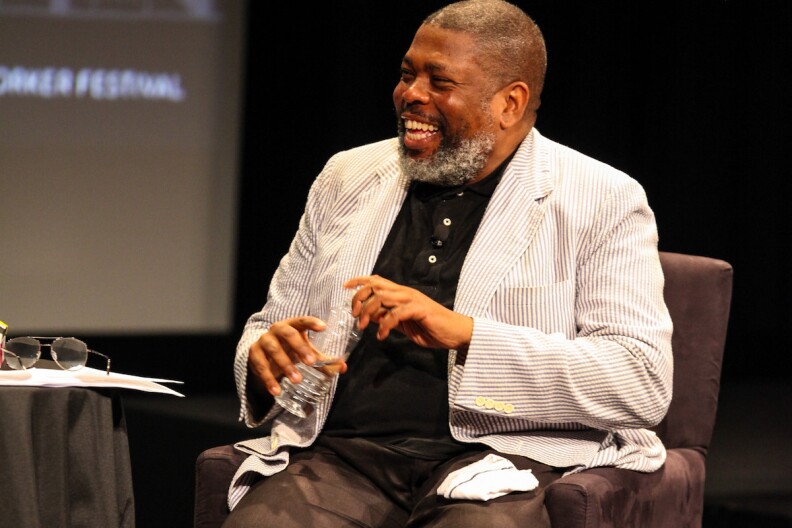 Hilton Als sits on a chair smiling with a bottle of water in his hand.