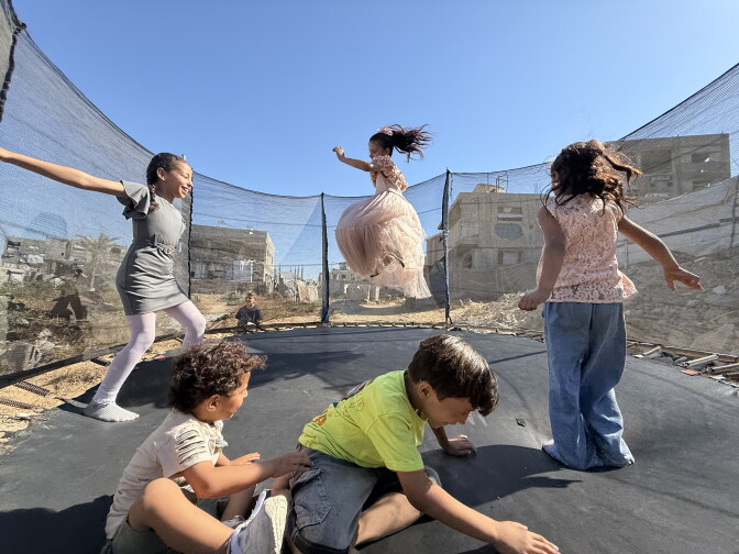 A group of five children play on a trampoline with mesh walls surrounding the trampoline. Three of them are jumping while two are seated. Surrounding the trampoline is rubble and some partially standing buildings.