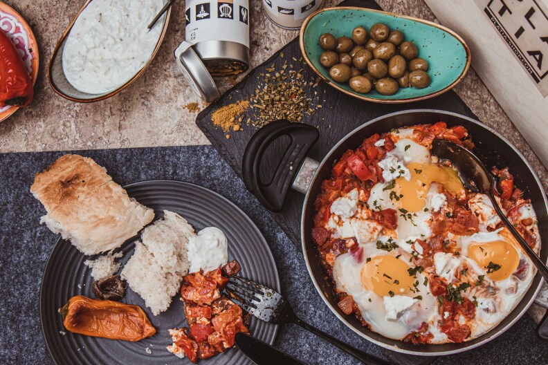 A photo of shakshuka in a pan, a bowl of olives, and then bread and roasted peppers on a plate.
