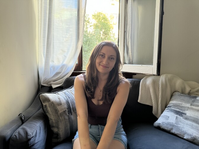 A woman with long brown hair looks at the camera and smiles slightly while sitting on a blue couch. Behind her is an open window.