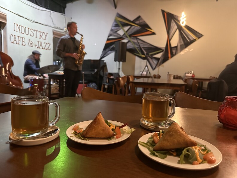 Food and drink sit on a table with a man playing saxophone in the background