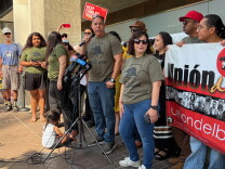 A man with medium skin tone speaks into a microphone. In front of him, there is a stand with several other microphones, each with a mic flag representing a  news organizations. About a dozen people stand around the man in the center, many wearing forest green shirts that read "UNION DEL BARRIO." On the ground, there is a small child playing with toy cars. 