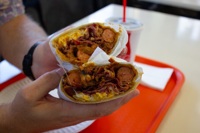 A pair of light-skinned hands holds a burrito-shaped food item cut in half, revealing the contents: two sliced pink hot dogs with thin ribbons of cooked red meat mixed with a thick brown chili sauce and shredded yellow cheese. 
