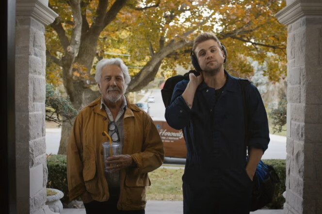 Two male presenting people, one elderly and one younger wearing headphones both with light skintones, stand outside the entrance to a house