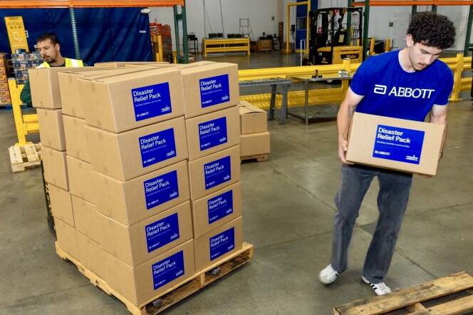 A male presenting person carries a cardboard box. A stack of about 30 boxes is next to him.