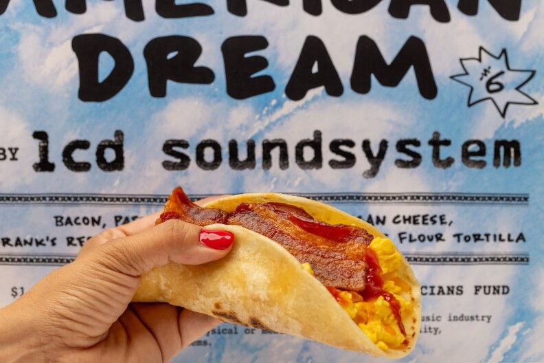 A female hand with red nail polish holds a taco with egg and bacon. In the background is a blue and white poster advertising the taco, including the "American Dream" and its price — $6.