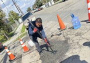A man with a medium dark skin tone stands on a street outside leaning down to stomp the ground with a tool. The tool is flat at the bottom to compact the aphalt that was freshly laid down over a damaged road area.