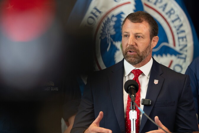 Homeland Security Secretary Markwayne Mullin, a man with light skin tone, wearing a dark blue suit and red tie, speaks into a microphone. Something out of focus in the foreground is partially in the left side of the frame.