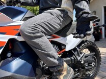 A close-up of a man on a blue, orange, and white motorcycle from the waist down. He's wearing brown boots that are resting on the bike's pegs, gray jeans, and a black and white leather jacket. His left arm is outstretched, with his left palm slightly raised and facing forward. 