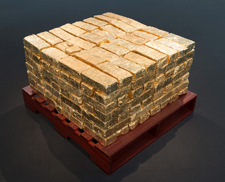 A pile of gold-painted bricks sits on a dark red colored palate on a gray-painted floor in front of a white wall.