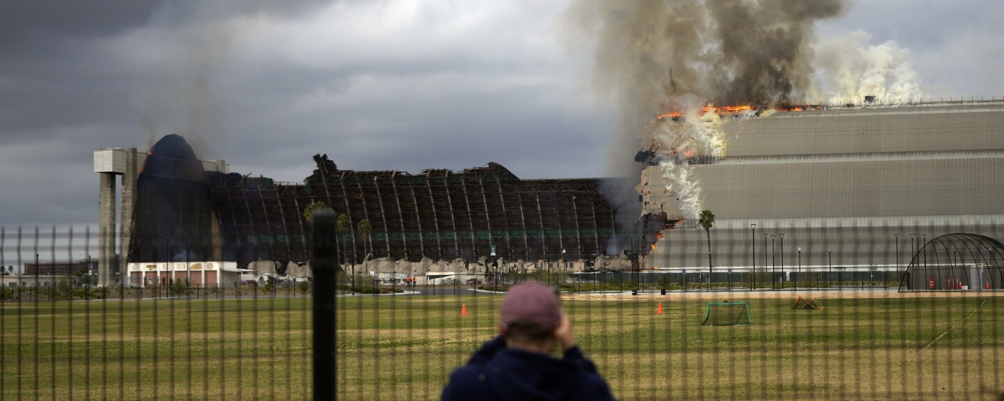 A person looks through a fence at a massive structure on fire, with at least half already destroyed by the flames. Dark smoke is billowing up into the air.