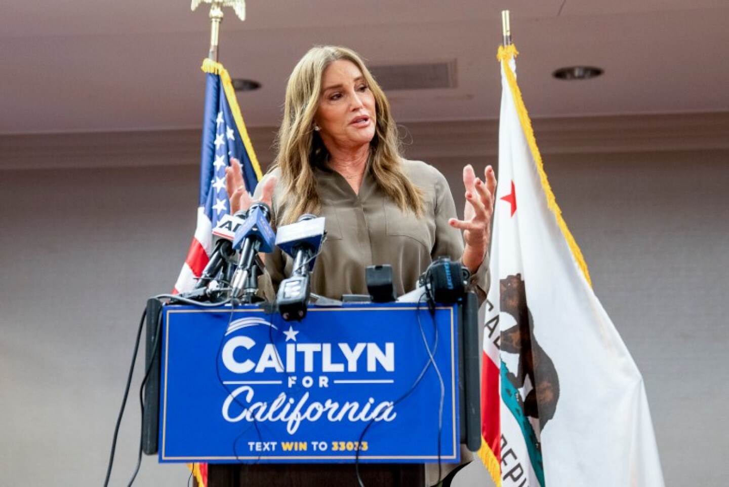Caitlyn Jenner stands at a lectern with a blue sign that says in white letters: Caitlyn for California. She has a state flag to her left and a U.S. flag to her right.