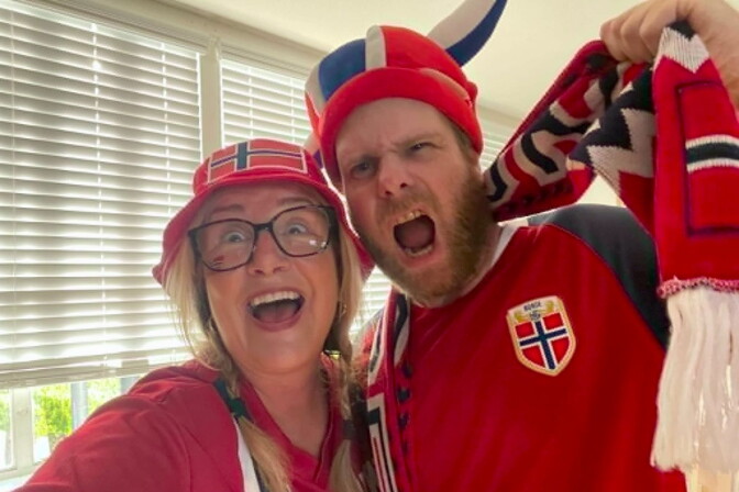 A woman and man with light skin tone both have their mouths open in excitement. The man wears a red, white and blue hat with Viking horns and the woman's red bucket hat has a flag with a dark blue cross in the left corner. 