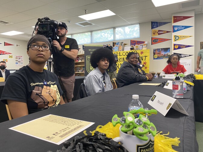 Four students sit at long tables lined with black tablecloths and gaze toward a screen that's out of frame behind the photographer. They have serious expressions on their faces. A television news camera points over the photographer's head in the direction of the screen.