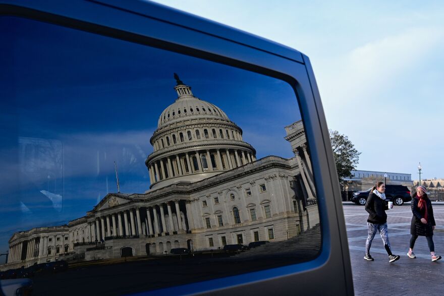 A reflection of the Capitol building in a dark car window during the day. People walk by in the background.