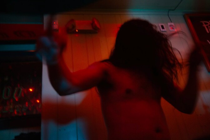 A man with long hair and no shirt on dances against a wood-paneled wall.