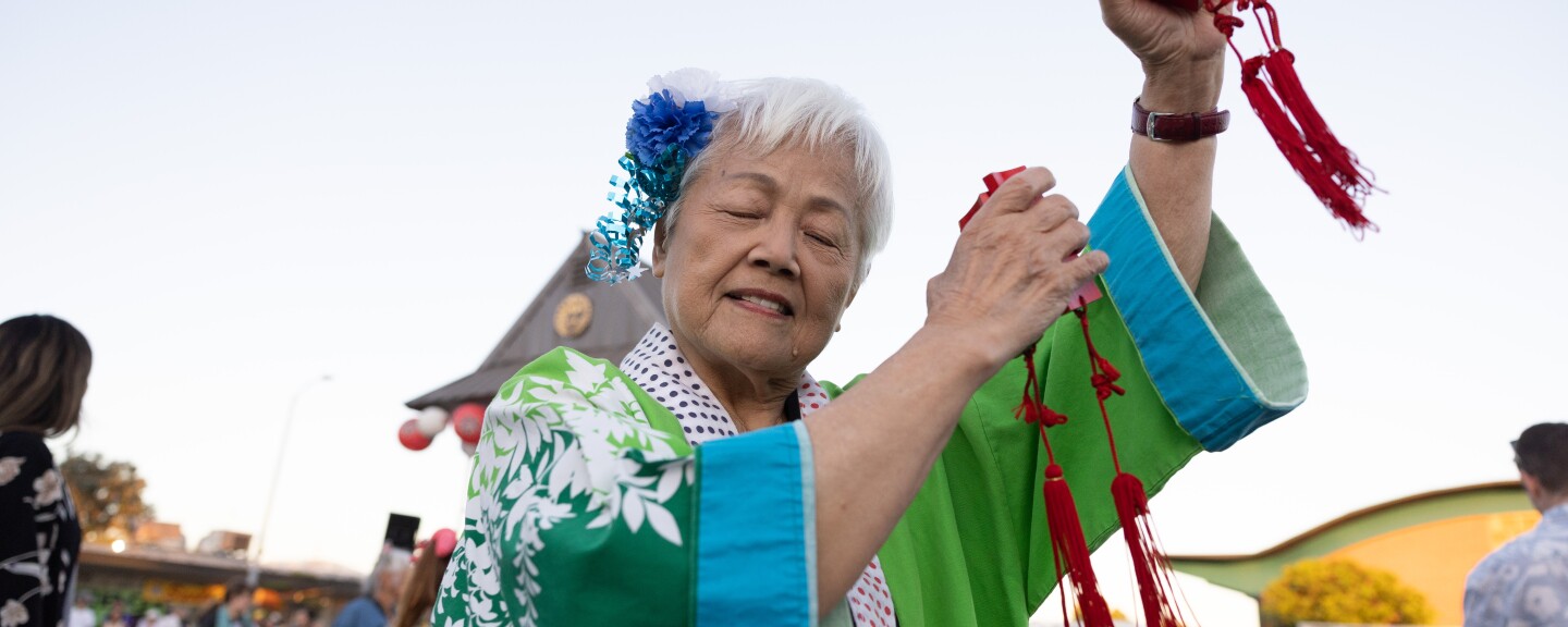 An Asian American woman with short white hair closes her eyes with her hands up in the air, holding red kachi kachi, or wooden castanets, with long red strings.