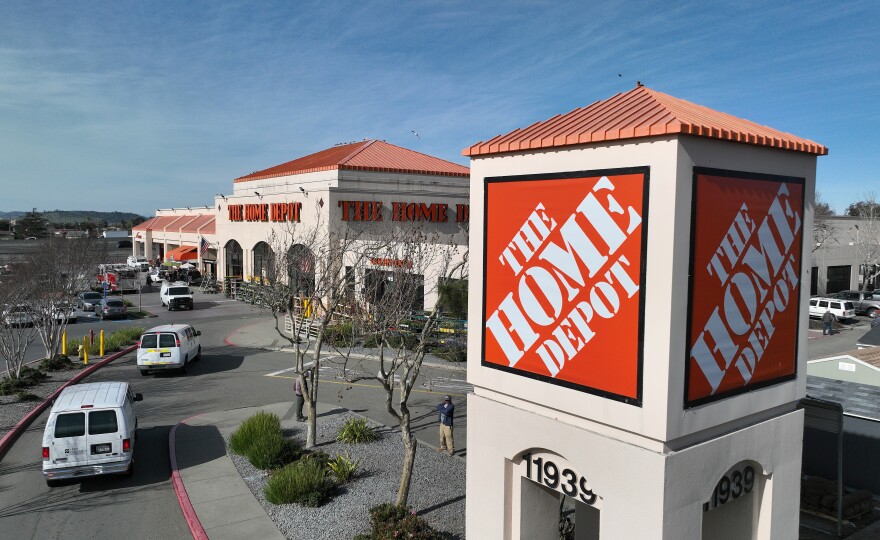 A aerial shot shows the outside of a Home Depot store. The sign for Home Depot is at the forefront of the image, white block letters against an orange background. To the left, white vans are moving to the front of the store entrance.