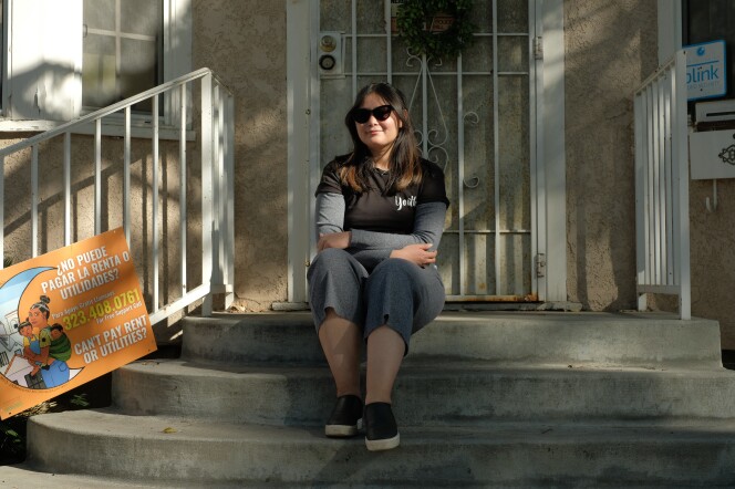 A young woman wearing a black t-shirt and grey pants sits on concrete steps leading to the front door of her house. 