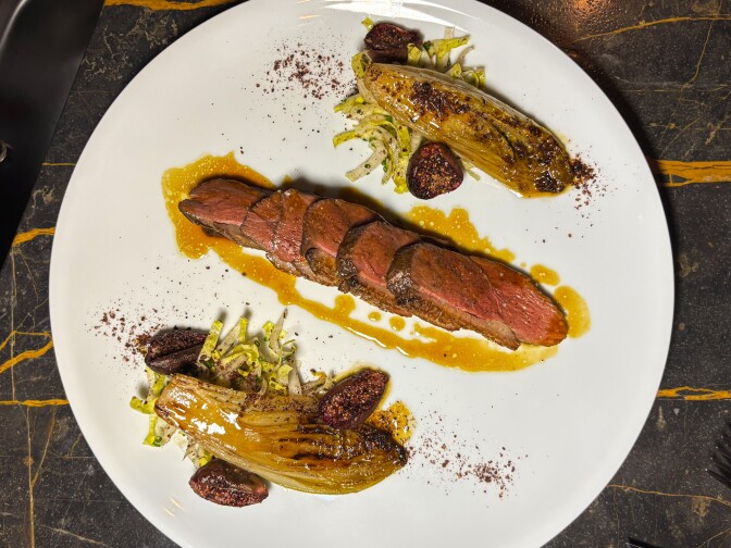 White plate with sliced duck breast in center showing pink interior, flanked by caramelized endive halves and roasted black figs, drizzled with golden sauce.
