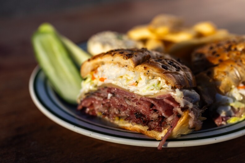 A close-up photo of a sandwich that has been cut in half and positioned to show off the inside: The sandwich has been made on an onion bread roll, and bit of poppy seed and charred onion speckles the top. The interior of the sandwich is made up of slices of dark, reddish pink colored meat that is piled high with a mound of coleslaw, with bits of orange shreds peeking through. There are also green pickle spears, a small serving of potato salad and golden brown French fries on the plate.