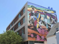 A tall affordable-housing building with a mural covering its upper side wall. The mural depicts Korean drummers in traditional dress mid-performance beneath sweeping ribbons of color, with green mountains and faint architecture in the background and large pink plum blossoms across the lower portion. Green trees and a peaked rooftop are in the foreground under a clear blue sky.