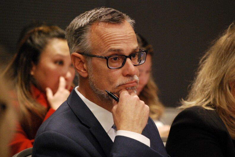 A man with light-tone skin wears a suit jacket and glasses and looks ahead while holding his hand to his chin, as other people sit around him.