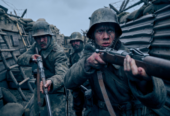 Felix Kammerer stands to the far right. He holds a rifle against his right shoulder ready to fire it if needed. Kammerer looks straightforward, eyes wide and intense, concentrating on what is in front of him. He wears a helmet and grey military uniform. His face is smudged with mud, a thin line of blood trailing along his nose. Behind Kammerer to the left is Albrecht Schuch. He too holds a rifle against his right shoulder, but its muzzle is pointed down towards the ground. His helmet hoods his eyes in shadow, and he wears the same grey military uniform as Kammerer. Behind them stands another man in the same helmet and uniform, his head and shoulders just visible between Kammerer's and Schuch's shoulders. The three men are surrounded by tall wooden and metal walls of war trenches. Everything is a grey hue. 