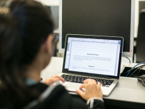 A person is sitting behind a laptop and typing with the screen reading "Application Essay".
