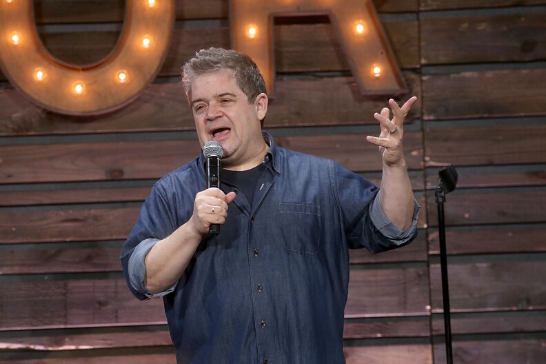 Patton Oswalt speaks into a microphone while performing on stage. He's wearing a blue denim shirt. Behind him, the wall has wood paneling and two metal letters in a partially visible sign are lit up.