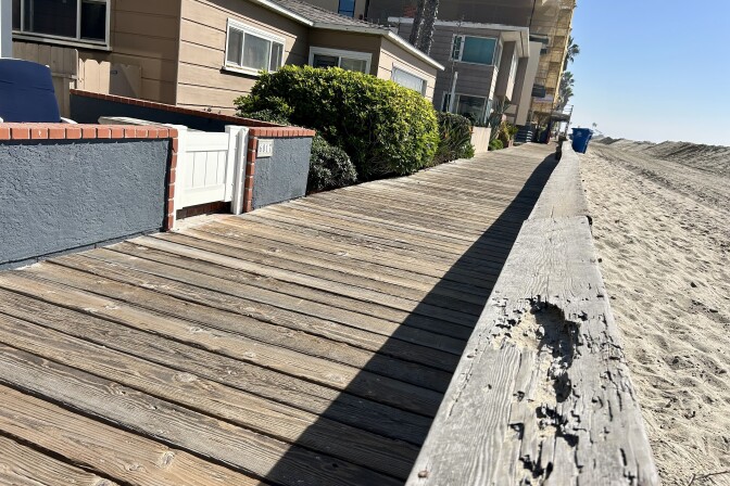 A wood walkway. Houses on the left and beach sand on the right of it.
