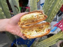 A light-skinned hand holds two halves of a sandwich stacked on top of each other. The sandwich contains ground brown meat mixed with cooked potatoes, shredded pale green lettuce, and crumbled white cheese, all inside a round bun. In the background, there is a large green succulent plant.