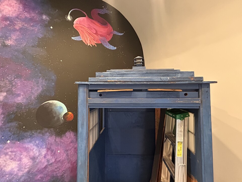 The top half of a navy blue telephone box. The box is missing its doors and has a ladder stowed inside.