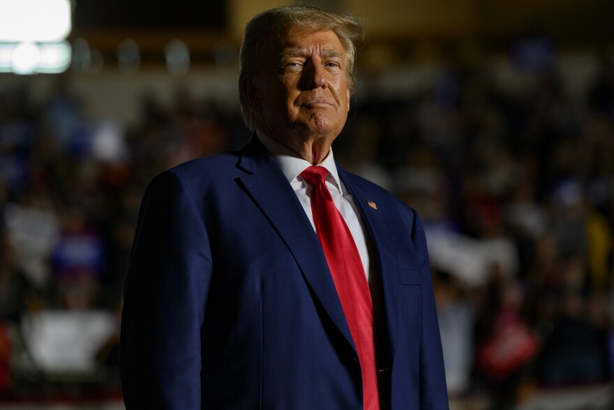 A man wears a dark suit coat and wide red tie. His hair is light and in a comb over.