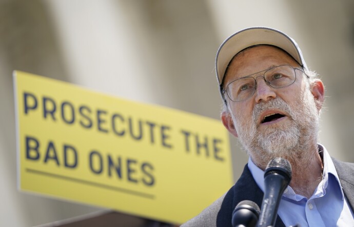A man wearing a baseball cap, eyeglasses and a dark blue suit jacket stands, speaking into a microphone. Behind him is a sign that reads, "prosecute the bad ones"