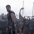 Actor Matt Damon stands on a gloomy shore wearing dark Greek armor. Tarps and historically Greek boats can be seen in the background.