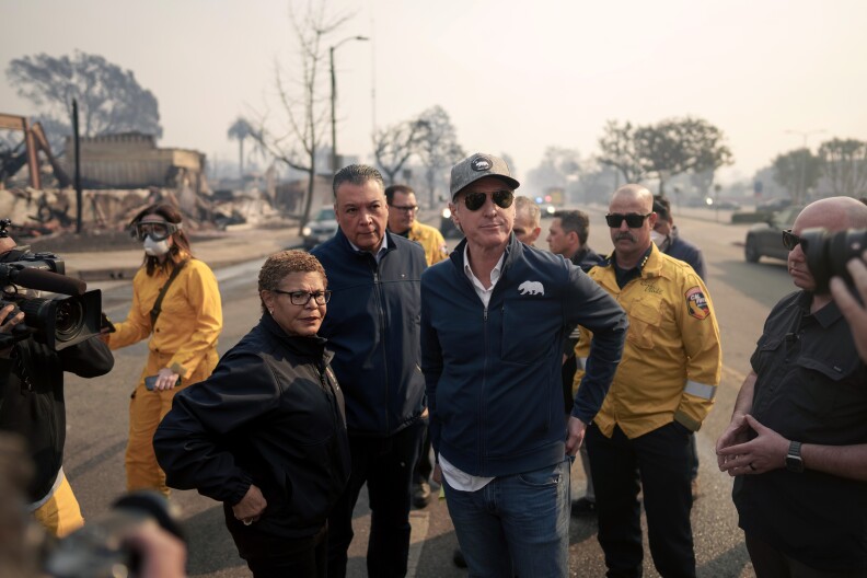 A man with light skin tone wearing a baseball cap and blue sweater with an icon of a bear stands next to a woman with medium-dark skin tone and short curly hair wearing glasses and black sweater. Behind them are various fire officials and reporters. In the background rubble from a fire can be seen.
