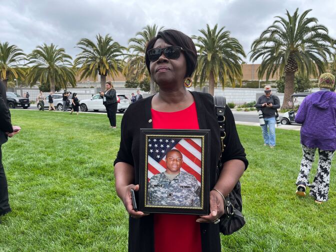 A woman with dark skin tone wearing a black sweater, red shirt and sunglasses hold a framed picture of a man in uniform, with dark skin tone and a U.S. flag in the background.
