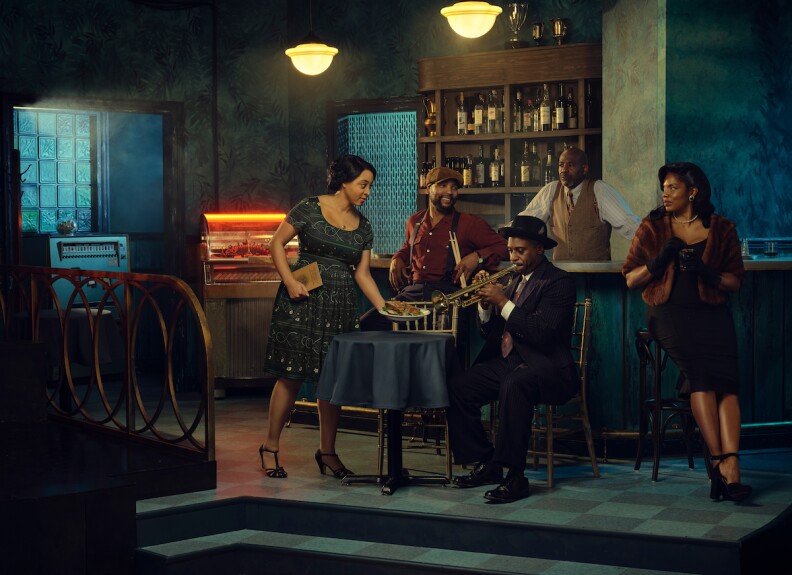 four people gather around a man sitting at a table and playing trumpet. everyone wears old timey clothes that look like they come from the 1930s or 40s