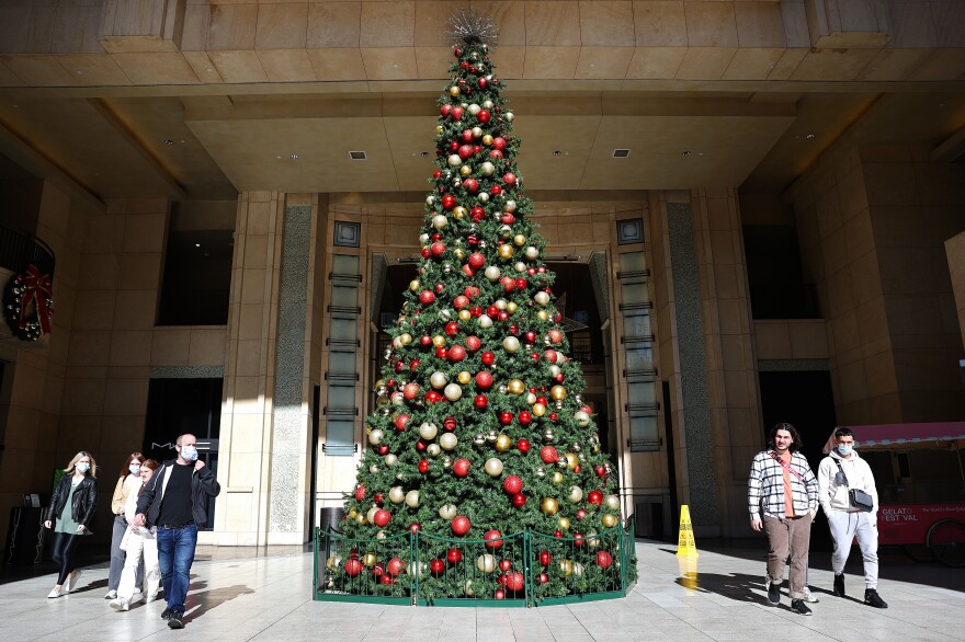 A massive Christmas tree decorated in gold and red ornaments sits in the entrance to a building . People walk past it on both sides, some masked, some not.
