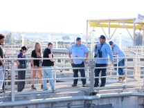 A group of high school students hear from adult water professionals in light blue attire at a water treatment facility outside on a sunny day. 