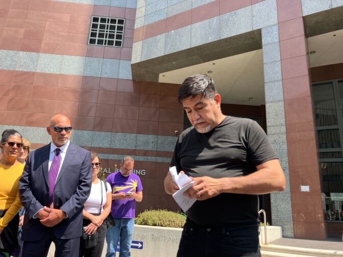 A Latino man in a black T-shirt and pants opens a piece of papaer while other look on outside a building.