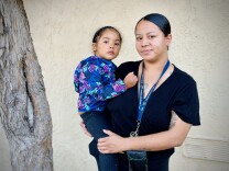  A medium skin-toned woman in a black top and a young medium skin-toned girl in a brightly patterned jacket stand together, the woman holding the girl close.