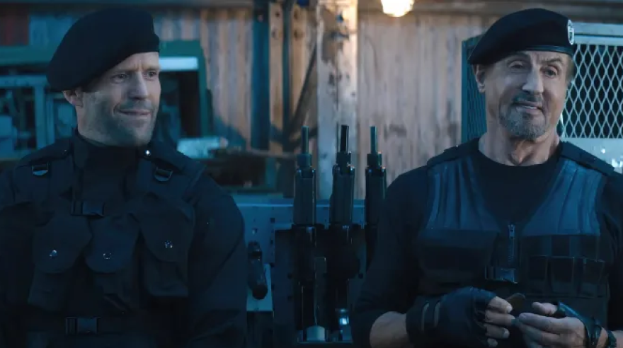 Jason Statham and Sylvester Stallone stand next to each other in black military gear, a rack of guns behind them. They are both smiling.