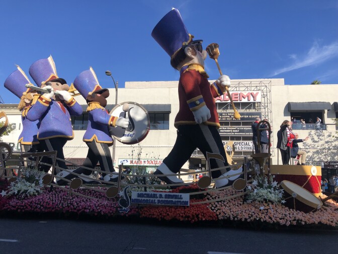 A float is covered in roses and carries a marching band leader figure, followed by figures playing the drums, flute and trombone. A sign reads: "The Michael D. Sewell Memorial Foundation."