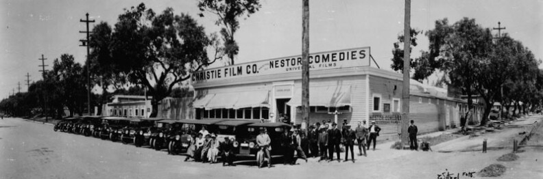 The first motion picture studio in Hollywood was built by David Horsley for Christie Film Co. Automobiles are lined up at Sunset Blvd., with Gower Street at right. The sign above the building reads "Christie Film Co.", and "Nestor Comedies, Universal Films." The photo includes Charles Christie, Horace Davey, Al Christie, Anton Nagy, head cameraman; George French, Gus Alexander, Harry Rattenburg, Lee Moran, Eddie Lyons, Billie Rhodes, and Eddie Barry. The photograph is signed "Witzel." 