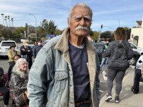 Richard Castaneda stands and waits for his mail outside the Pasadena Post Office on Lincoln Ave. His home burned down in the 2025 Eaton Fire. He wares a weathered jean jacket. He has a bushy white mustache. 