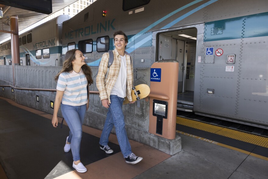 A female presenting person wearing blue jeans and a striped t-shirt walks alongside a male presenting person carrying a skateboard wearing blue jeans, a white t-shirt and a open, button down shirt. In the background, the doors to an open Metrolink train are visible.