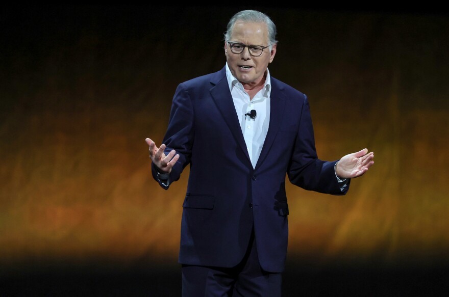 A man who appears to be white and in his 50s stands on a stage speaking, wearing a navy suit and white shirt.