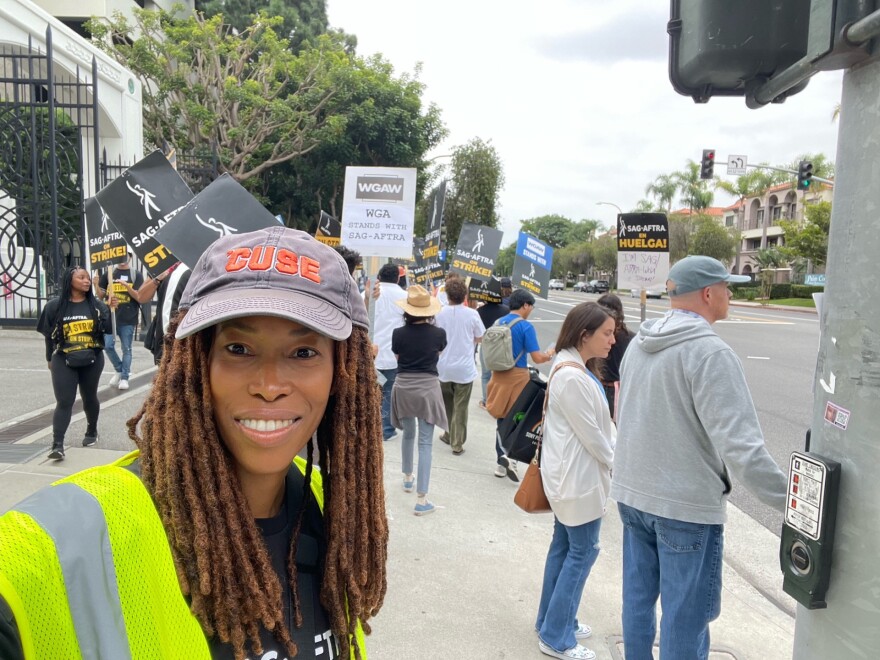 A dark-skinned woman in a ball cap and bright neon vest looks toward the camera smiling. Picketing writers and actors can be seen on the sidewalk behind her holding signs. 